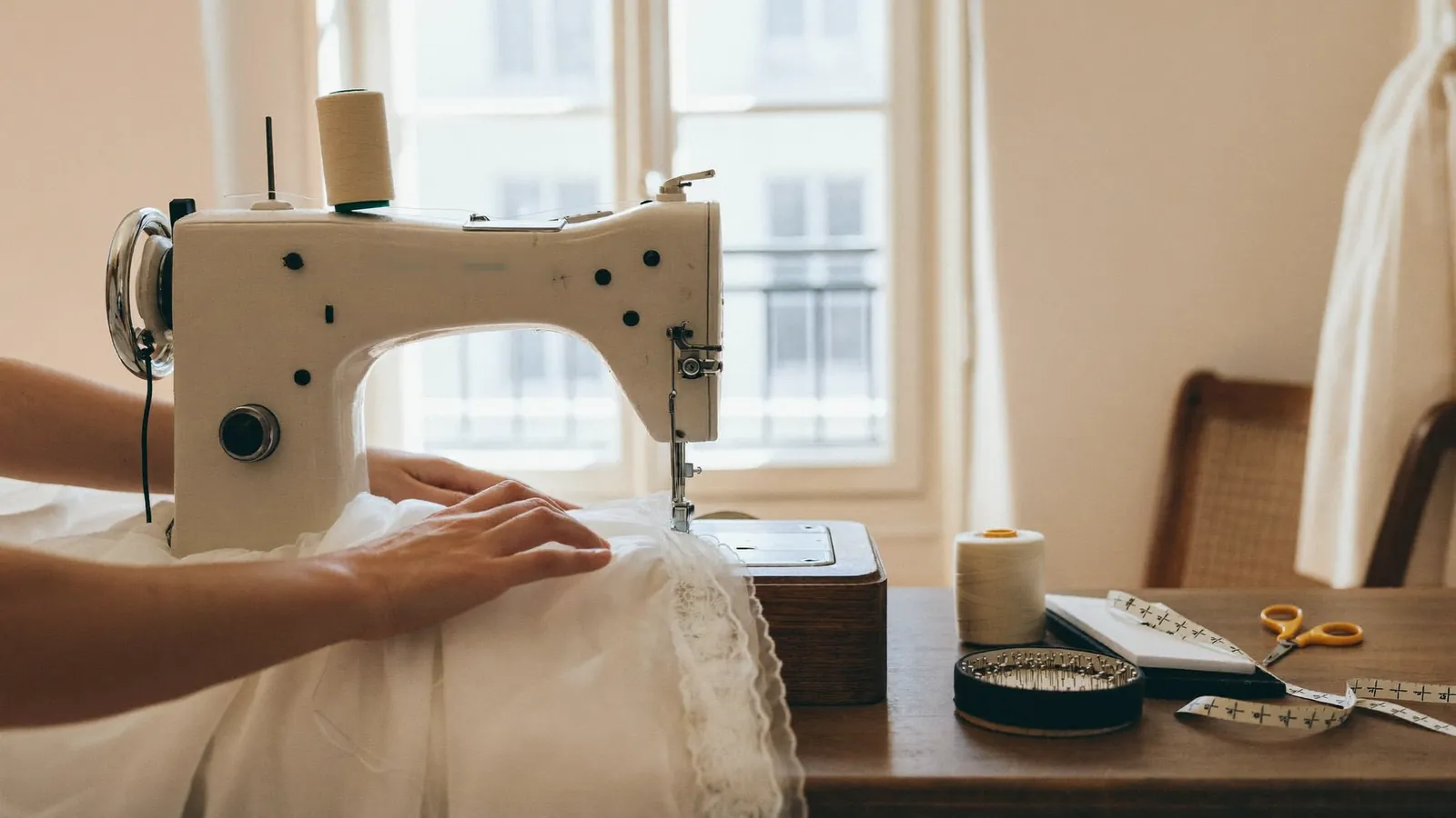 Hände an der Nähmaschine mit weißem Brautstoff im hellen Atelier, Maßband und Stecknadeln auf dem Tisch