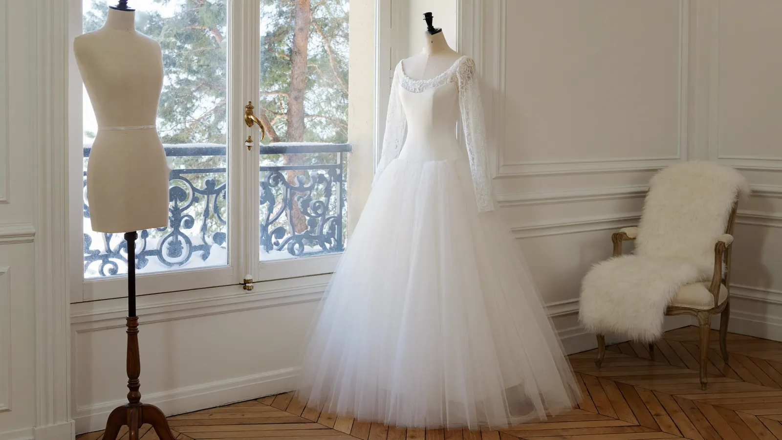 Brautkleid mit langen Spitzenärmeln und Tüllrock an einer Schneiderpuppe am Fenster, verschneiter Garten draußen, Pelz-Sessel im hellen Winter-Atelier